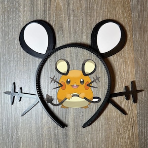 Puede incluir: Una diadema negra con orejas de ratón adjuntas y una imagen de dibujos animados de un personaje Dedenne. Las orejas son negras con secciones internas blancas. La imagen de Dedenne es naranja, amarilla y blanca.