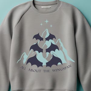 Puede incluir: Sudadera gris con un gráfico de murciélagos volando sobre una cordillera con el texto "All About The Wingspan".