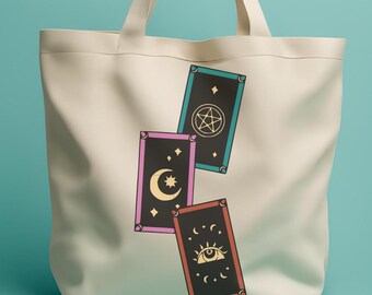 May include: A white canvas tote bag with three tarot cards printed on it. The cards are in a stack, with the top card showing a pentagram, the middle card showing a crescent moon and star, and the bottom card showing an eye.