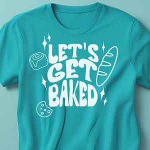 May include: A turquoise blue t-shirt with the text "Let's Get Baked" in white. The text is in a retro font style. There are images of a cinnamon roll, a baguette, and a cookie around the text.