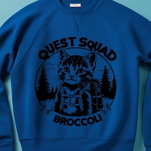 Op de afbeelding: Een blauw sweatshirt met een zwarte afbeelding van een kat met een rugzak en de tekst "Quest Squad Broccoli".