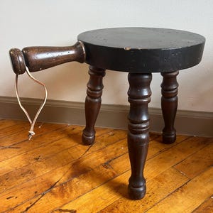 May include: A dark brown wooden stool with a round top and three turned legs. The stool has a single wooden handle attached to the side, with a leather strap hanging from it.