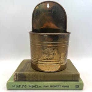 May include: A vintage brass wall pocket with a decorative sun face, resting on two antique books. The pocket has a curved top and a cylindrical body with horizontal bands. The books are green and have the title "MEATLESS MEALS".