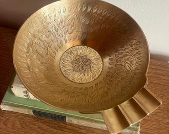 May include: A vintage brass ashtray with an intricate floral design. The ashtray has a wide, shallow bowl and a curved, ridged lip.