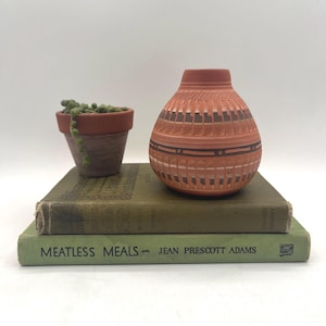 May include: A terracotta-colored, patterned vase sits atop two vintage green books, one titled "Meatless Meals." A small terracotta pot with a trailing succulent is to the left. The vase has a geometric design with dark brown and light brown accents.