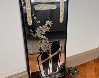 May include: A rectangular black framed mirror with a decorative design. The design includes a glittery gold shape at the top, a floral arrangement, and two vases. The mirror is leaning against a white wall and brown floor.