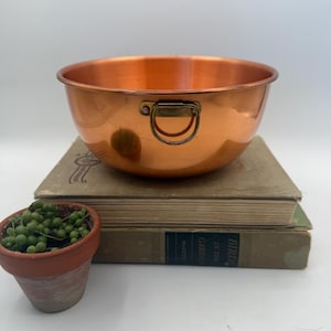 May include: A polished copper mixing bowl with a rounded base and a single brass ring handle attached to the side. The bowl features a smooth, metallic finish and a wide, open top, reflecting light across its curved, warm-toned surface.