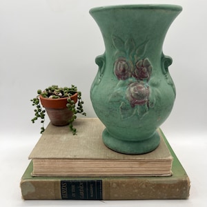May include: A vintage green vase with embossed floral details and two small handles. The vase is displayed on top of two antique books, one green and one tan. A small potted plant with trailing vines is to the left.