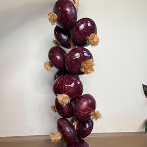 May include: A string of decorative, deep purple garlic bulbs with tan-colored, fibrous stems. The bulbs are strung together with a thick, natural-colored rope. The garlic is hanging against a white wall.