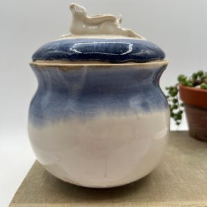May include: A ceramic jar with a lid, featuring a gradient of blue and white. The lid is topped with a cream-colored sculptural element. The jar is sitting on a vintage book, suggesting a decorative or storage item.