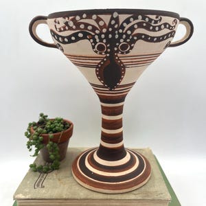 May include: A tall, decorative ceramic chalice with a wide rim and two handles. The chalice features a brown and white octopus design on the bowl and a striped stem. The base is round and striped. A small potted plant and books are in the background.