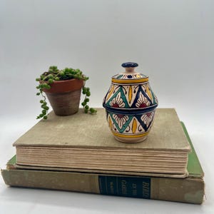 May include: A small, decorative ceramic jar with a lid, featuring a colorful geometric pattern in yellow, teal, navy, and red. The jar sits atop two vintage books, with a small potted plant to the left.