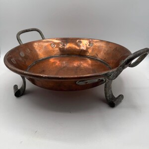 May include: A round copper serving dish with a polished metallic finish, featuring two curved iron handles on opposite sides and three sturdy, scrolled iron legs attached to the base. The interior surface shows a warm, reddish-brown hue.