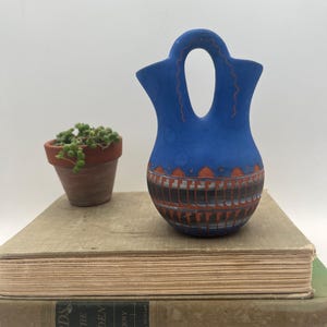 May include: A blue ceramic vase with a handle, decorated with orange and black geometric patterns. The vase is displayed on a stack of old books, next to a small terracotta pot with a green plant.
