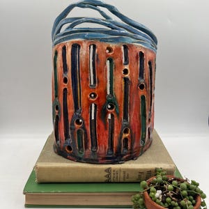 May include: A cylindrical ceramic lantern with a blue handle and rim, featuring vertical cutouts and a vibrant color palette of red, green, and navy. The lantern sits atop two vintage books, with a small potted plant in the foreground.