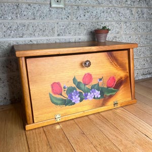 May include: A rectangular wooden bread box with a drop-down front door featuring a hand-painted floral design of red tulips and purple violets. The door has a clear glass knob and two brass-colored metal hinges at the base for opening.