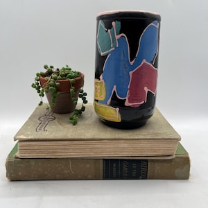 May include: Black cylindrical vase with abstract blue, pink, and yellow shapes. The rim is pink. The vase sits on two vintage books, next to a small terracotta pot with a trailing plant.