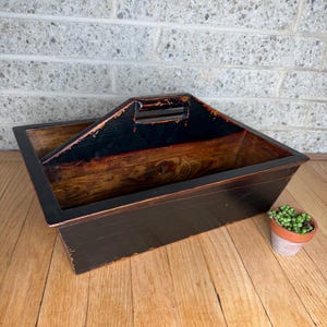 May include: A rectangular wooden caddy with a central handle, featuring a distressed black exterior finish and a natural stained wood interior. The edges show signs of wear, revealing the wood grain beneath the dark paint on the handle.