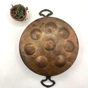 May include: A vintage copper escargot pan with seven circular indentations. The pan has two ornate handles and a dark patina. A small terracotta pot with a trailing plant is in the upper left corner.