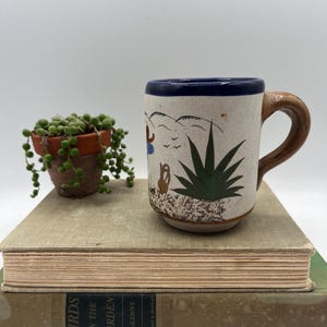 May include: A ceramic mug with a brown handle and a blue rim. The mug features a hand-painted design of a desert landscape with an agave plant and figures. A small potted plant sits next to the mug on top of a stack of books.