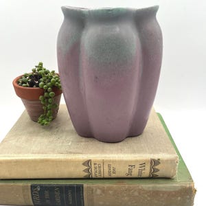 May include: A two-toned, lavender and green ceramic vase with a scalloped rim. A small terracotta pot with a trailing succulent sits to the left of the vase. The vase is on top of two vintage books.