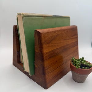 May include: A wooden book stand with a smooth, polished finish and visible grain, holding a single hardcover book with a green cover. Next to the stand is a small terracotta pot containing a green succulent plant with round, bead-like leaves.