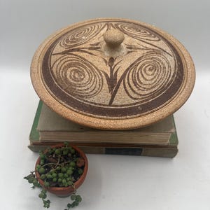 May include: A tan ceramic lidded dish with a brown spiral design. The lid has a small knob and sits atop a stack of old books. A small terracotta pot with a trailing plant is in the foreground.