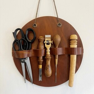 May include: A round, brown leather tool holder with a variety of leatherworking tools. The tools include black scissors, a metal edge creaser, a wooden burnisher, a metal awl, and a wooden mallet. The holder has a leather strap to secure the tools and a hanging cord.
