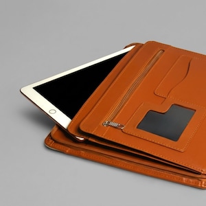 Può includere: Una cartella in pelle marrone chiaro con chiusura a cerniera e molteplici tasche interne. Un tablet bianco con schermo nero è parzialmente inserito nella cartella. La cartella ha una tasca trasparente e uno scomparto con cerniera.