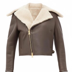 Puede incluir: Chaqueta de cuero marrón con forro y cuello de borrego color crema. La chaqueta tiene una cremallera dorada y un diseño corto. Estilo aviador clásico.