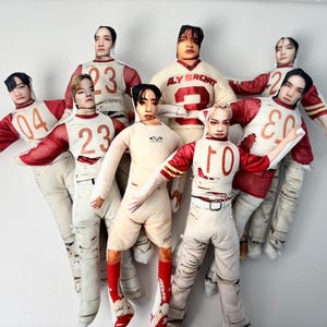 May include: A group of seven fabric dolls, each with a human face and wearing a sports jersey. The jerseys are primarily white with red sleeves and accents, and feature numbers. One doll wears a cream-colored jersey with the text "LY SPORT".