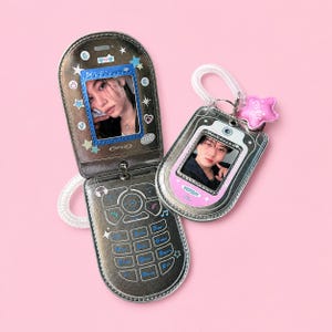 May include: A metallic dark grey flip phone-shaped photo holder with a blue frame displaying a person's portrait and a keypad graphic. A smaller matching metallic grey photo holder with a pink frame and a pink star charm is attached by a clear coil.