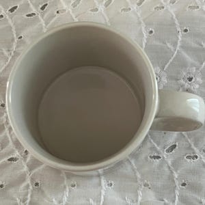 May include: A plain, off-white ceramic mug with a smooth, cylindrical interior and a sturdy, curved handle attached to the side. The mug features a simple, minimalist design with a wide, open top, showcasing a clean, uniform finish throughout.