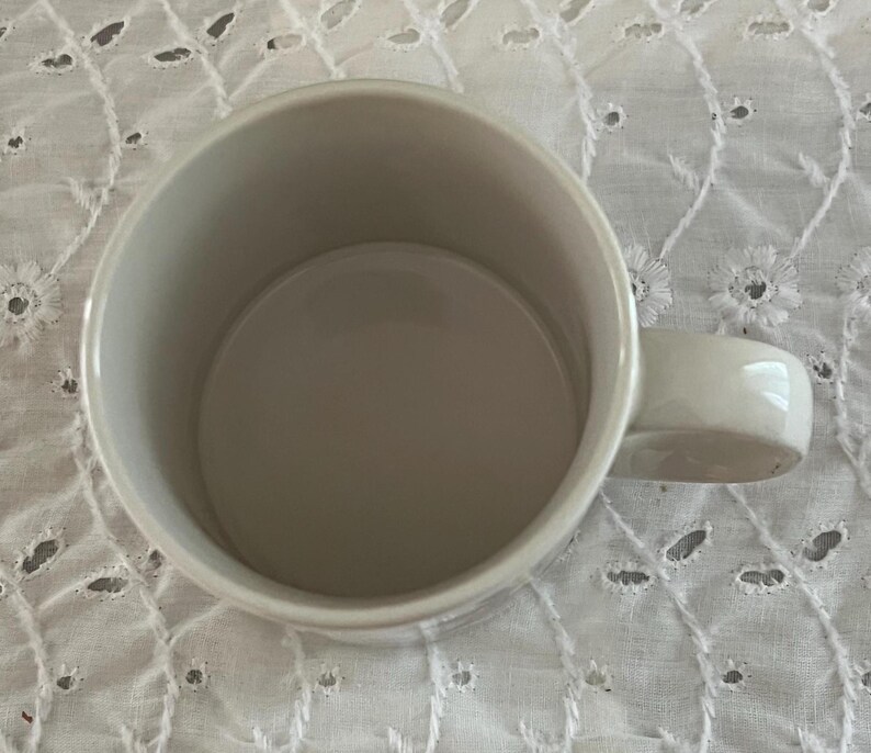 May include: A plain, off-white ceramic mug with a smooth, cylindrical interior and a sturdy, curved handle attached to the side. The mug features a simple, minimalist design with a wide, open top, showcasing a clean, uniform finish throughout.