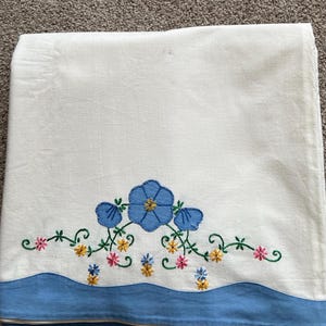 May include: White linen towel with a blue floral embroidery design. The embroidery features a large blue flower with yellow accents, surrounded by smaller flowers in pink and yellow, and green vines. A blue border is at the bottom.