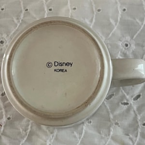 May include: The underside of a white ceramic mug featuring a circular base with a handle extending to the right. Printed in black text in the center is the copyright symbol followed by the word Disney and the word KOREA underneath it.