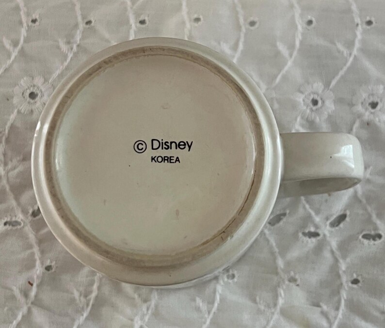 May include: The underside of a white ceramic mug featuring a circular base with a handle extending to the right. Printed in black text in the center is the copyright symbol followed by the word Disney and the word KOREA underneath it.