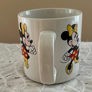 May include: A white ceramic mug featuring a repeating graphic print of Minnie Mouse. The character is depicted with a yellow bow, a red dress with yellow polka dots, and yellow shoes. The mug has a smooth, glossy finish and a curved handle on the side.