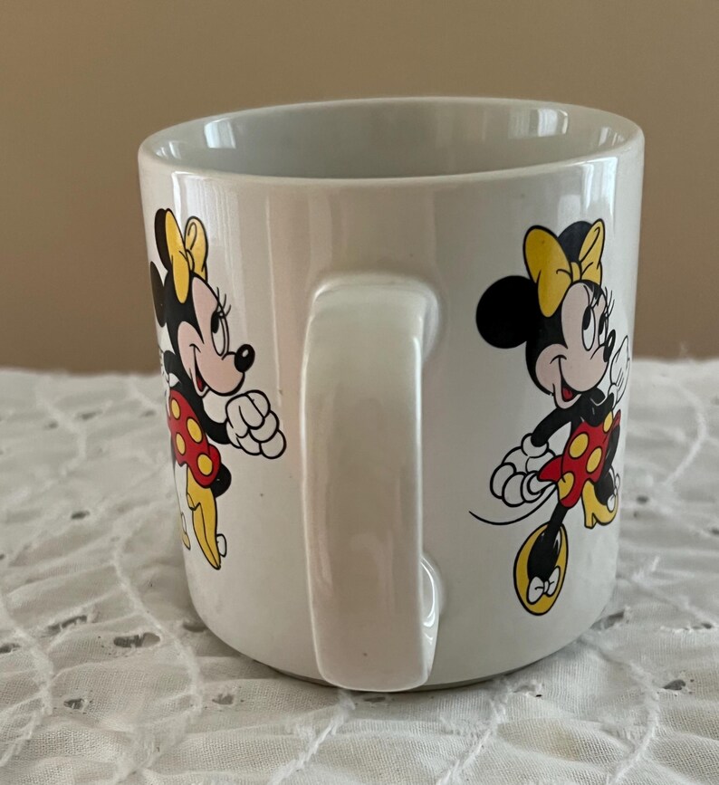 May include: A white ceramic mug featuring a repeating graphic print of Minnie Mouse. The character is depicted with a yellow bow, a red dress with yellow polka dots, and yellow shoes. The mug has a smooth, glossy finish and a curved handle on the side.