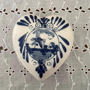 May include: A heart-shaped ceramic trinket box with a blue and white Delft design. The box features a windmill, a house, and a boat in a circular design.