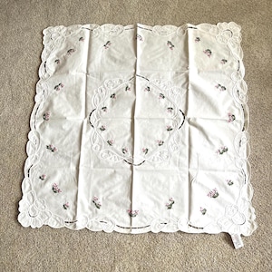 May include: A square off-white tablecloth featuring a central circular embroidered pattern and a scalloped border with cutwork details. The fabric is decorated with small pink floral motifs and green leaves scattered across the surface.