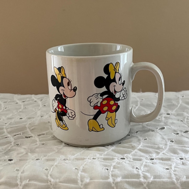 May include: A white ceramic mug featuring two illustrations of Minnie Mouse. One image shows her standing and looking to the left, while the other shows her walking to the right. She wears a yellow bow, a red skirt with yellow polka dots, and yellow shoes.