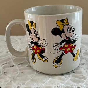 May include: A white ceramic mug featuring a repeating graphic print of Minnie Mouse. The character is depicted wearing a yellow bow, a red skirt with yellow polka dots, and yellow shoes. The mug has a curved handle on the left side and a smooth finish.