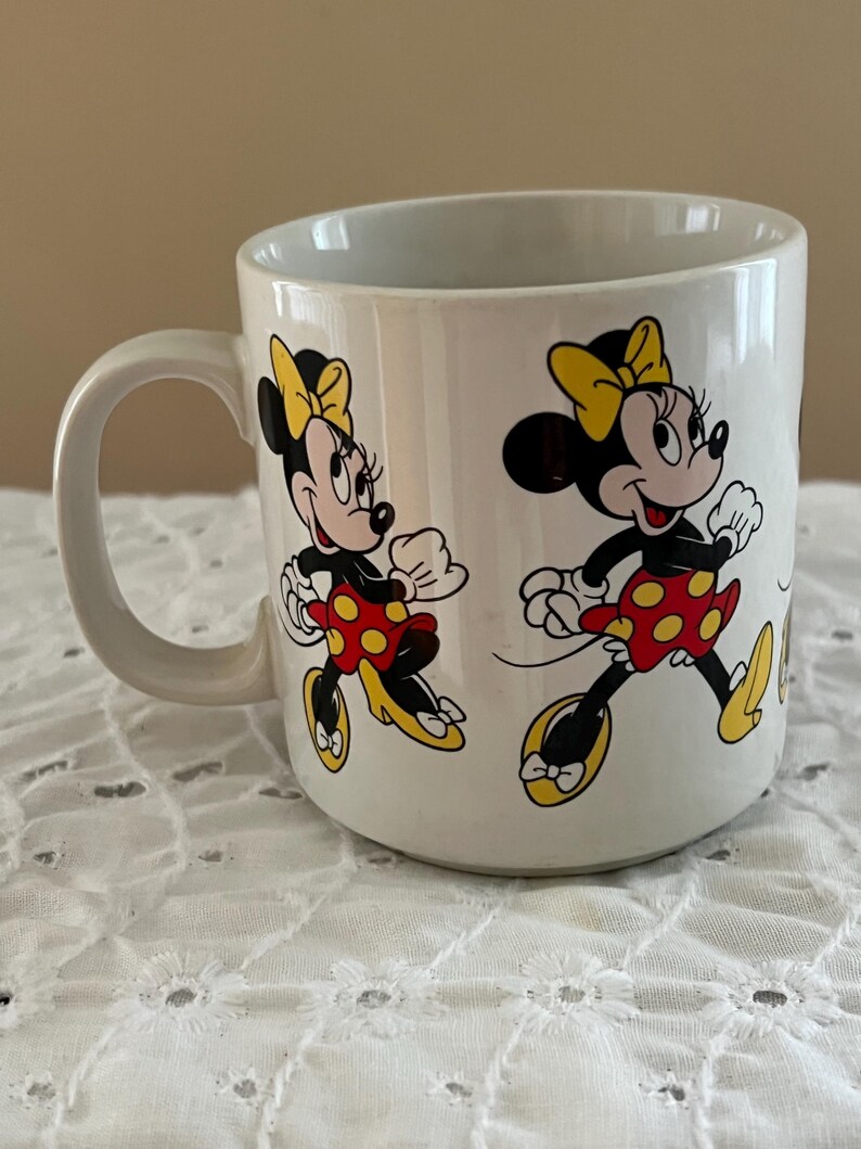 May include: A white ceramic mug featuring a repeating graphic print of Minnie Mouse. The character is depicted wearing a yellow bow, a red skirt with yellow polka dots, and yellow shoes. The mug has a curved handle on the left side and a smooth finish.