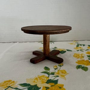 May include: A small, round, dark brown wooden pedestal stand featuring a flat circular top surface supported by a single central pillar and a cross-shaped base. The object is constructed from stained wood with a visible grain texture throughout.