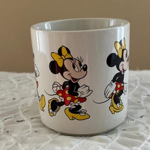 May include: A white ceramic mug featuring a repeating graphic print of Minnie Mouse. The character is depicted in various poses, wearing a yellow bow, a red skirt with yellow polka dots, and yellow high-heeled shoes. The mug has a smooth finish.