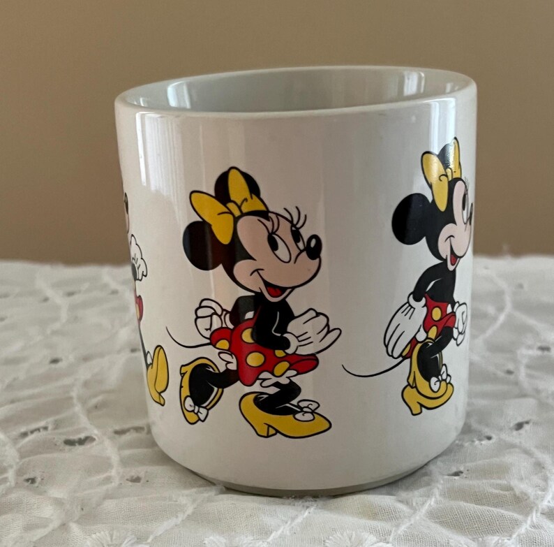 May include: A white ceramic mug featuring a repeating graphic print of Minnie Mouse. The character is depicted in various poses, wearing a yellow bow, a red skirt with yellow polka dots, and yellow high-heeled shoes. The mug has a smooth finish.