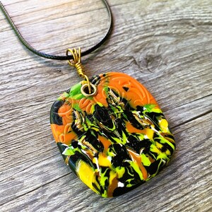 May include: A black cord necklace with a gold wire wrapped around a square pendant. The pendant is made of resin and has a swirling pattern of black, orange, yellow, and green.