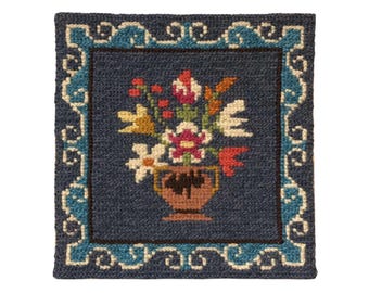 Op de afbeelding: Een blauw tapijt met een bloemenmotief. Het tapijt heeft een vaas met bloemen in tinten roze, rood, geel en wit. De vaas is bruin en heeft een witte rand. Het tapijt heeft een blauwe rand met een wit en blauw patroon.