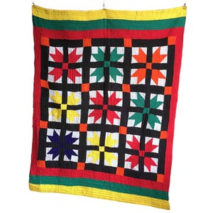 Puede incluir: Un edredón colorido con un patrón de estrellas geométricas. El edredón presenta una cuadrícula negra con formas de estrella rojas, verdes, naranjas, amarillas y azules. El borde es rojo, con rayas verdes y amarillas.